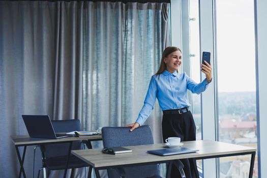 Image of a happy young woman in a jacket smiling and working on a laptop, talking on the phone in a modern office with large windows. Remote work