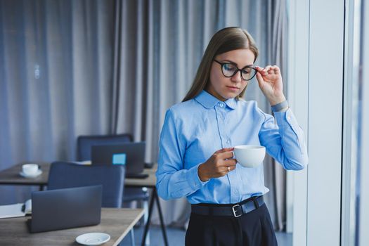 Image of a happy young woman in a classic shirt and glasses smiling and drinking coffee while standing by the window in a modern office with large windows. Remote work