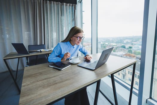 Adult smiling female manager in glasses and formal shirt sits at a desk and looks at a laptop. Modern, spacious, bright office with a large window.