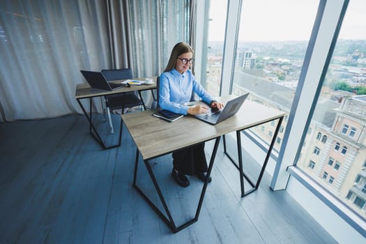 Adult smiling female manager in glasses and formal shirt sits at a desk and looks at a laptop. Modern, spacious, bright office with a large window.