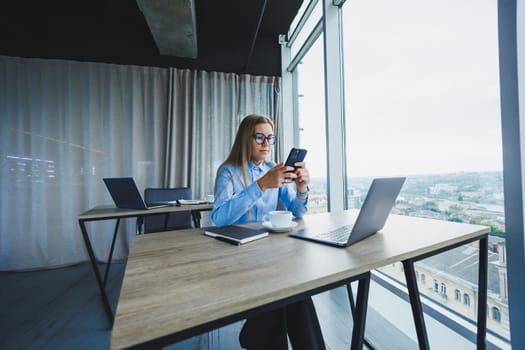 Adult smiling female manager in glasses and formal shirt sits at a desk and looks at a laptop. Modern, spacious, bright office with a large window.