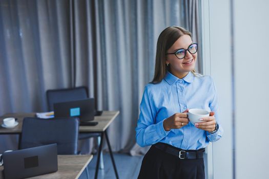 Image of a happy young woman in a classic shirt and glasses smiling and drinking coffee while standing by the window in a modern office with large windows. Remote work