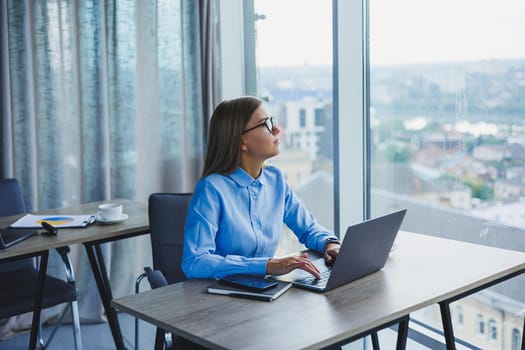 Young beautiful business woman in a blue shirt sitting in the office with a laptop and doing business