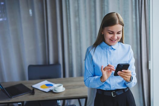 Image of a happy young woman in a jacket smiling and working on a laptop, talking on the phone in a modern office with large windows. Remote work