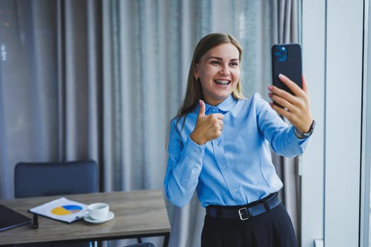 Image of a happy young woman in a jacket smiling and working on a laptop, talking on the phone in a modern office with large windows. Remote work