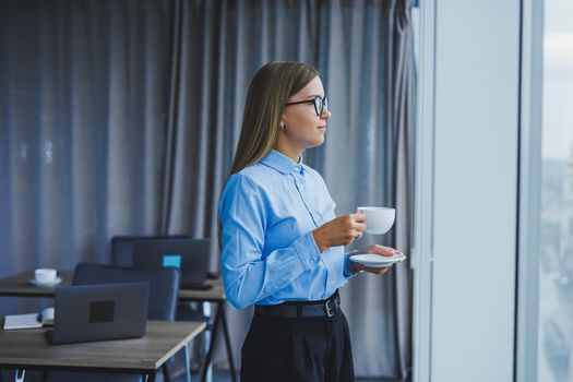 Image of a happy young woman in a classic shirt and glasses smiling and drinking coffee while standing by the window in a modern office with large windows. Remote work