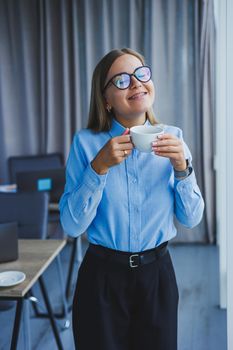 Image of a happy young woman in a classic shirt and glasses smiling and drinking coffee while standing by the window in a modern office with large windows. Remote work