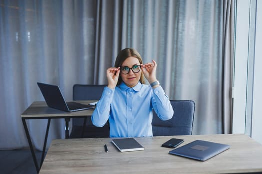 A beautiful woman in glasses sits at a table in the office and works at a laptop. Corporate work in the office. Modern woman workplace manager in classic clothes