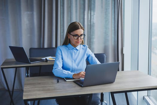 A beautiful woman in glasses sits at a table in the office and works at a laptop. Corporate work in the office. Modern woman workplace manager in classic clothes