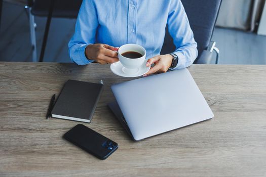 Portrait of cheerful woman in classic glasses smiling in free time in cafe with coffee, positive european woman in blue shirt, desk with laptop, remote work