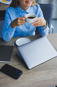 Portrait of cheerful woman in classic glasses smiling in free time in cafe with coffee, positive european woman in blue shirt, desk with laptop, remote work