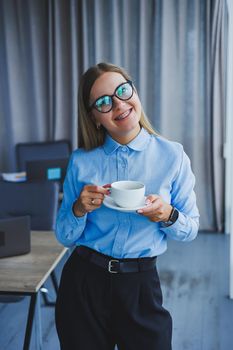 Image of a happy young woman in a classic shirt and glasses smiling and drinking coffee while standing by the window in a modern office with large windows. Remote work
