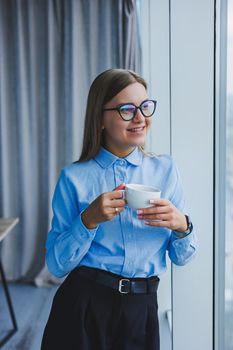 Image of a happy young woman in a classic shirt and glasses smiling and drinking coffee while standing by the window in a modern office with large windows. Remote work
