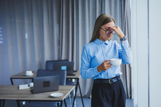 Image of a happy young woman in a classic shirt and glasses smiling and drinking coffee while standing by the window in a modern office with large windows. Remote work