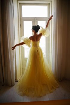 A woman's silhouette in a golden luxurious dress against the background of a window holds a curtain with her hands. Elegant lady in a yellow long silk dress with bare back, back view