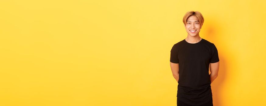 Portrait of friendly handsome asian guy with blond hair, smiling politely, holding hands behind back, standing yellow background.