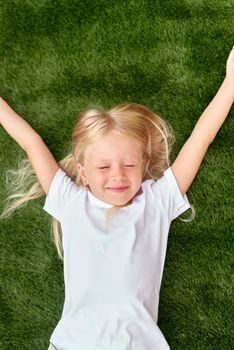 Top view. Mock up. Pretty beautiful blonde child girl lying and stretching on green grass. Smiling preschool girl 5-6 years old in white t shirt. Lifestyle Summer vacations Leisure People concept