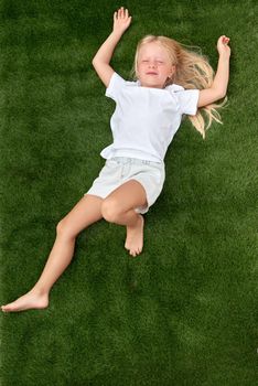 Top view. Mock up. Pretty beautiful blonde child girl lying and stretching on green grass. Smiling preschool girl 5-6 years old in white t shirt. Lifestyle Summer vacations Leisure. People concept