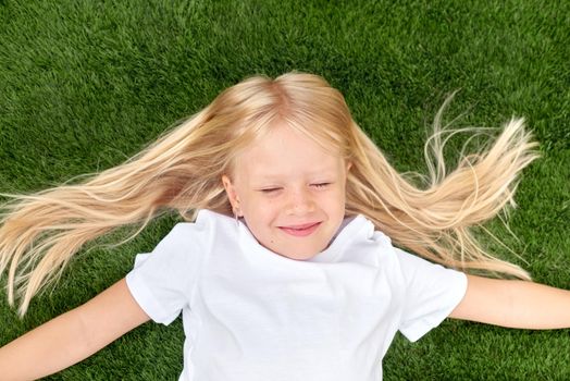 Top view. Mock up. Pretty beautiful blonde child girl lying and stretching on green grass. Preschool girl 5-6 years old in white t shirt. Lifestyle Summer vacations Leisure. People concept