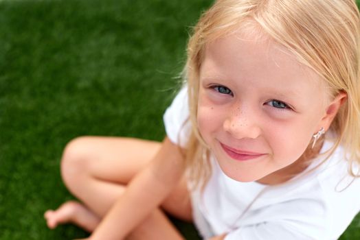 Top view. Pretty beautiful blonde child girl sitting on green grass. Smiling preschool girl 5 - 6 years old in white t shirt looking at camera. Lifestyle. Beauty. Summer vacations. leisure. people