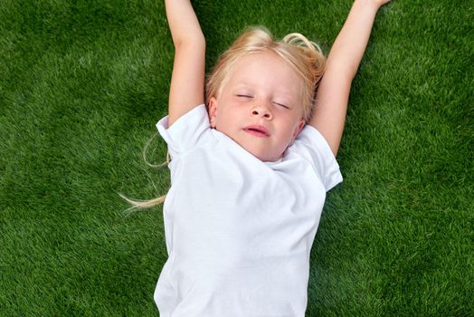 Top view. Mock up for logo, text, design. Blonde child girl closed eyes lying stretching on green grass. Preschool girl 5-6 years old in white t shirt. Lifestyle Summer vacations Leisure. People