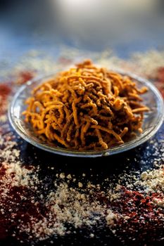 Diwali and Shravan Som special Teekha Gathiya in a glass plate along with some spread chickpea flour, red chili powder, and other ingredients that are needed to make the snack on a black surface.