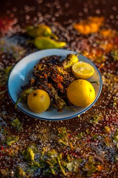 Close up shot of Classic Bharela Marcha or Stuffed fried chilies in a glass plate along with lemons and all the spices needed for preparing it on a brown wooden surface.