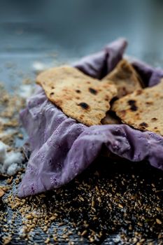 Close up shot of round bread Bhakri on the black wooden surface along with some raw whole wheat, salt in a container. Shot of Bhakri in a container on the black surface.