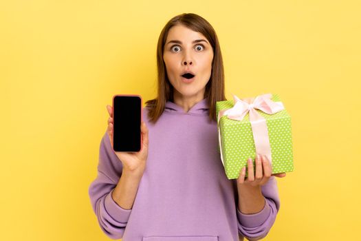 Present, bonus for mobile user. Portrait of amazed surprised woman holding gift box and cell phone with mock up, blank display, wearing hoodie. Indoor studio shot isolated on yellow background.