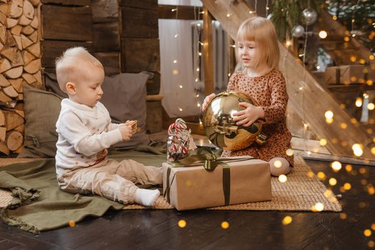 Little brother and sister play on Christmas eve in a beautiful house decorated for the New Year holidays. Children are playing with a Christmas gift. Scandinavian-style interior with live fir trees and a wooden staircase.