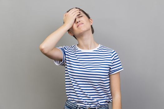 Portrait of upset frustrated woman wearing striped T-shirt holding forehead with hand, making facepalm gesture, forget about date, big failure. Indoor studio shot isolated on gray background.