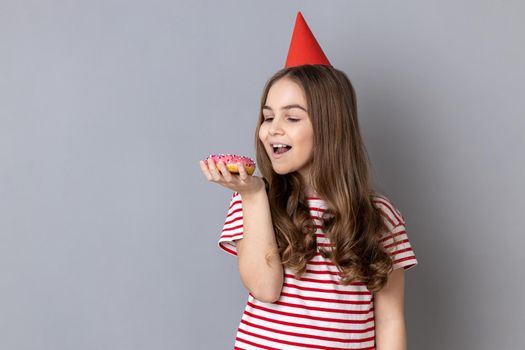 Portrait of hungry joyful little girl wearing striped T-shirt and party cone, holding and want to bite tasty sweet donut, looking at dessert. Indoor studio shot isolated on gray background.