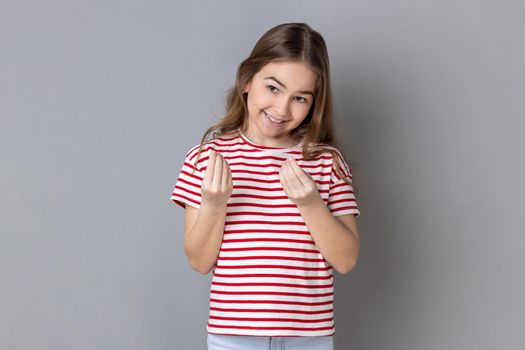 Portrait of smiling little girl wearing striped T-shirt makes money gesture, rubs fingers, looking at camera with glad expression. Indoor studio shot isolated on gray background.