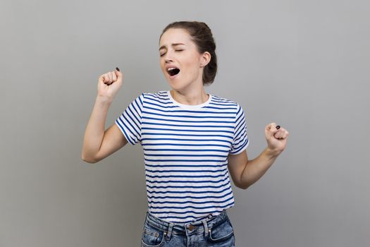 Portrait of tired sleepy woman wearing striped T-shirt yawning and raising hands up, feeling fatigued, standing with close eye. Indoor studio shot isolated on gray background.