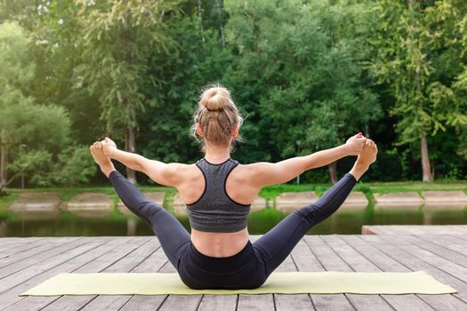 A slender woman sits on green mat, on a wooden platform by a pond in the park, in summer, does yoga. Copy space