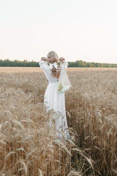 A blonde woman in a long white dress walks in a wheat field. The concept of a wedding and walking in nature.