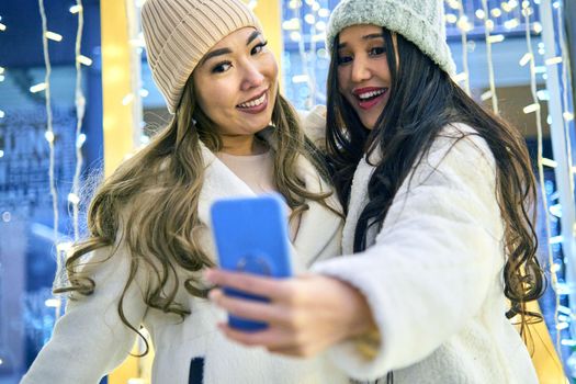 Two girlfriends asian girls in white coats are having fun hugging and selfie on mobile phone, new year, light bulbs
