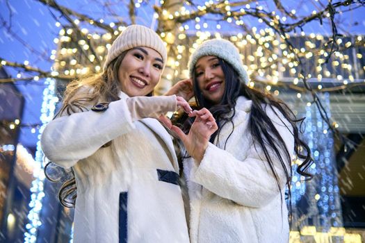 Two girlfriends asian girls in white coats are having fun, showing heart, hugging, new year, light bulbs