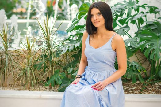 An outdoor portrait of an attractive brunette woman wearing a blue long dress is posing by the fountain at the park