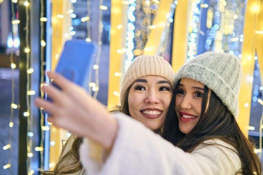 Two girlfriends asian girls in white coats are having fun hugging and selfie on mobile phone, new year, light bulbs