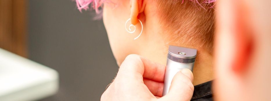 Back view of hairdresser's hand shaving nape and neck with electric trimmer of young caucasian woman with short pink hair in beauty salon