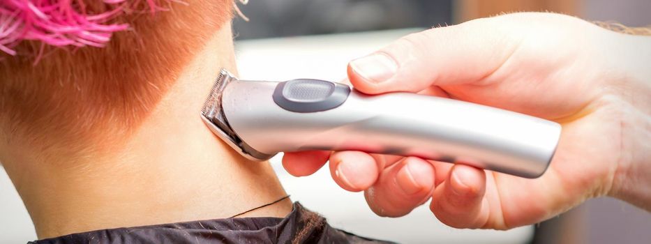 Back view of hairdresser's hand shaving nape and neck with electric trimmer of young caucasian woman with short pink hair in beauty salon