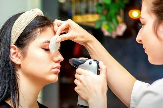 Side view of a make-up artist using aerograph making an airbrush makeup foundation on a female face in a beauty salon