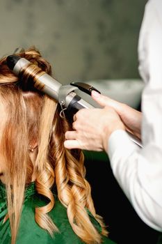 Hairdresser using curling tongs curls long brown hair on the young caucasian girl in a beauty salon