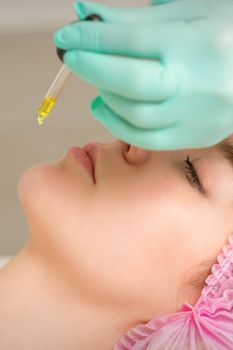 Close up of beautician holding pipette and applying oil serum drop on the face of a young woman in spa salon