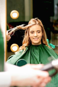 Hairdresser using curling tongs curls long brown hair on the young caucasian girl in a beauty salon