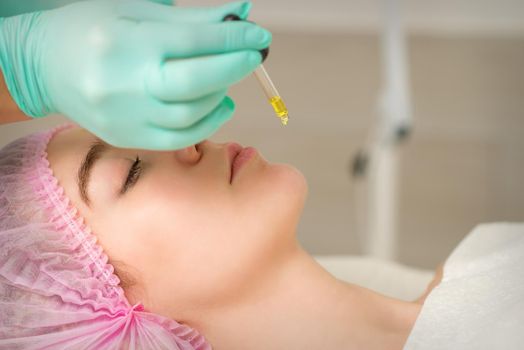 Close up of beautician holding pipette and applying oil serum drop on the face of a young woman in spa salon