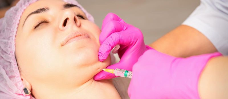 The cosmetologist makes lipolytic injection on the chin of a young woman against the double chin in a beauty salon