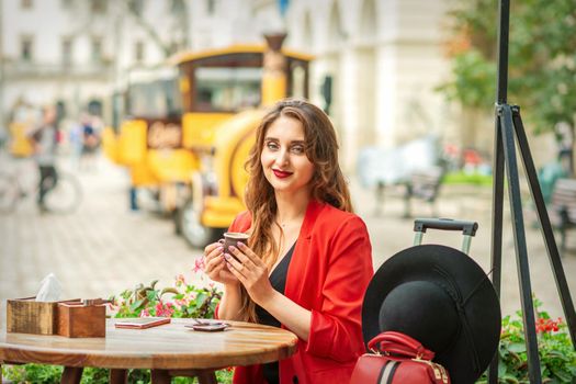 Tourist young caucasian woman in red jacket with coffee cup at the table in cafe outdoors