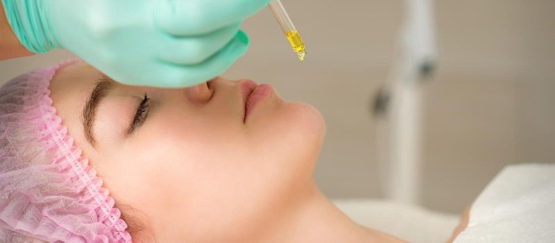 Close up of beautician holding pipette and applying oil serum drop on the face of a young woman in spa salon
